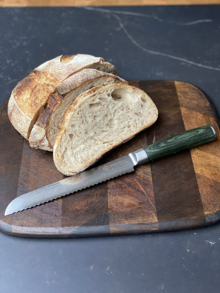 Een versgebakken brood met een knapperige korst en zachte binnenkant ligt op een houten snijplank. Ernaast ligt een HexClad broodmes met een groen handvat, klaar om het brood in perfecte sneetjes te snijden.