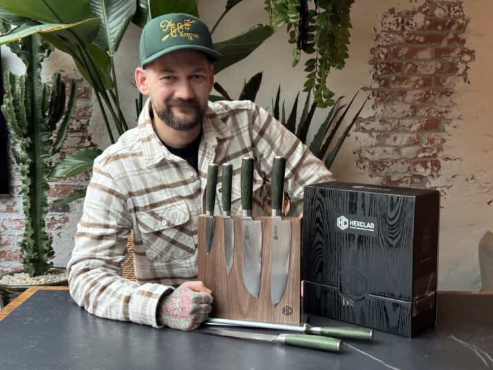 Jan Willem Huffmeijer poseert met een HexClad messenblok in een rustieke setting met bakstenen muren en veel groen. Hij draagt een groene pet en een geruite blouse, en leunt ontspannen op de tafel naast de luxe messenverpakking.