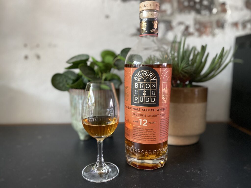 Een fles Berry Bros & Rudd 12 Year Old Speyside Sherry Cask whisky staat op een donkere tafel naast een gevuld whiskyglas. De diepe amberkleur van de whisky springt eruit, terwijl de groene planten en bakstenen muur op de achtergrond zorgen voor een warme en rustieke sfeer. De fles lijkt subtiel boven het oppervlak te zweven, wat een speels en uniek effect geeft aan de foto.