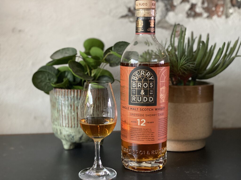 Een fles Berry Bros & Rudd 12 Year Old Speyside Sherry Cask whisky staat op een donkere tafel, vergezeld door een whiskyglas gevuld met de amberkleurige dram. Op de achtergrond staan groene kamerplanten in rustieke potten, wat de setting een warme en stijlvolle uitstraling geeft. De fles heeft een elegante label met koperkleurige accenten en benadrukt de rijke sherryvatrijping van deze Speyside single malt.