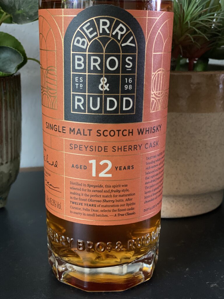 Een gedetailleerde close-up van het label op de fles Berry Bros & Rudd 12 Year Old Speyside Sherry Cask whisky. Het label toont de klassieke branding met koperkleurige accenten en beschrijft de rijping in Oloroso sherryvaten. De diepe amberkleur van de whisky schittert door de heldere fles, terwijl de achtergrond met groene planten en een rustieke setting de luxe uitstraling benadrukt.