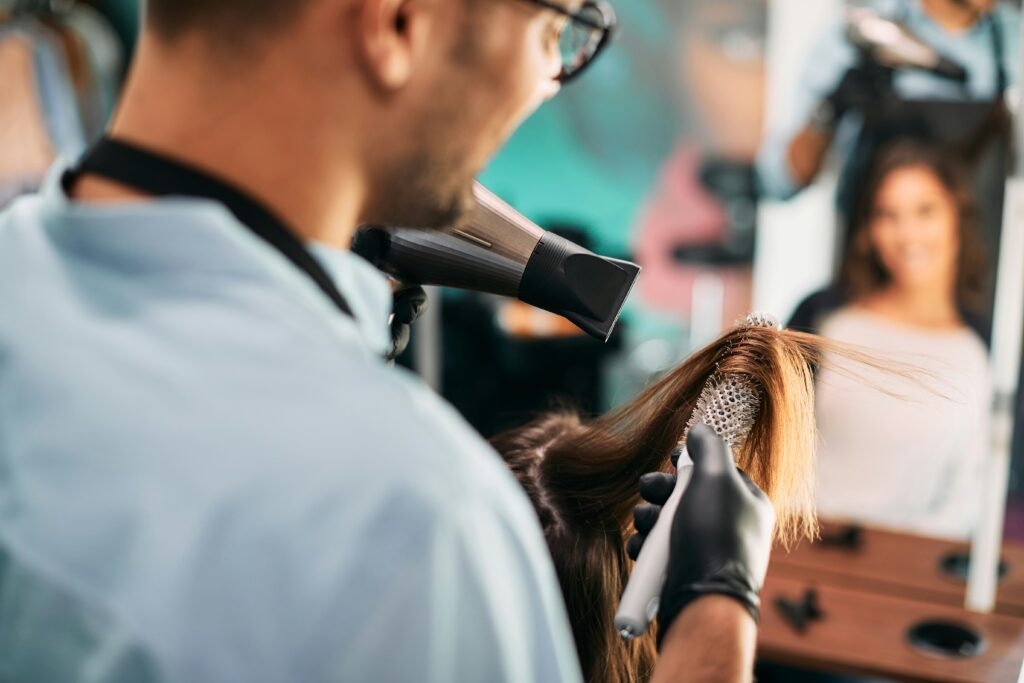 Een kapper met handschoenen gebruikt een föhn en een ronde borstel om het haar van een klant in een moderne kapsalon te stylen. In de spiegel is de klant zichtbaar, glimlachend naar het resultaat.