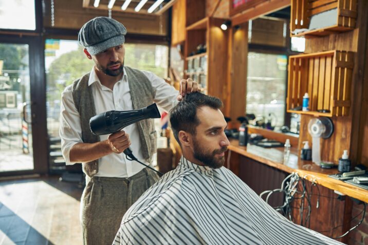 Een barbier met een klassieke outfit en een platte pet droogt het haar van een klant met een föhn in een traditioneel ingerichte barbershop. De klant draagt een gestreepte kapperscape en kijkt geconcentreerd vooruit.