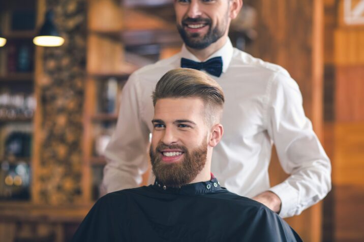 Glunderende man met perfect gestyled haar en een verzorgde baard bij de kapper, genietend van een frisse nieuwe look.